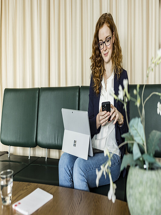 Eine Frau sitzt mit Computer und Smartphone auf dem Sofa und arbeitet in entspannter Atmosphäre